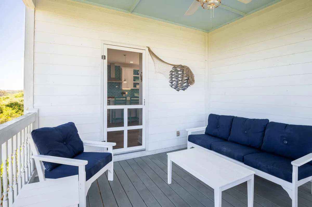 A breezy kitchen porch with white wicker furniture and blue cushions set on a wooden deck, accented by decorative netting. The porch provides an inviting spot for morning coffee or sunset cocktails with a hint of coastal ambiance.