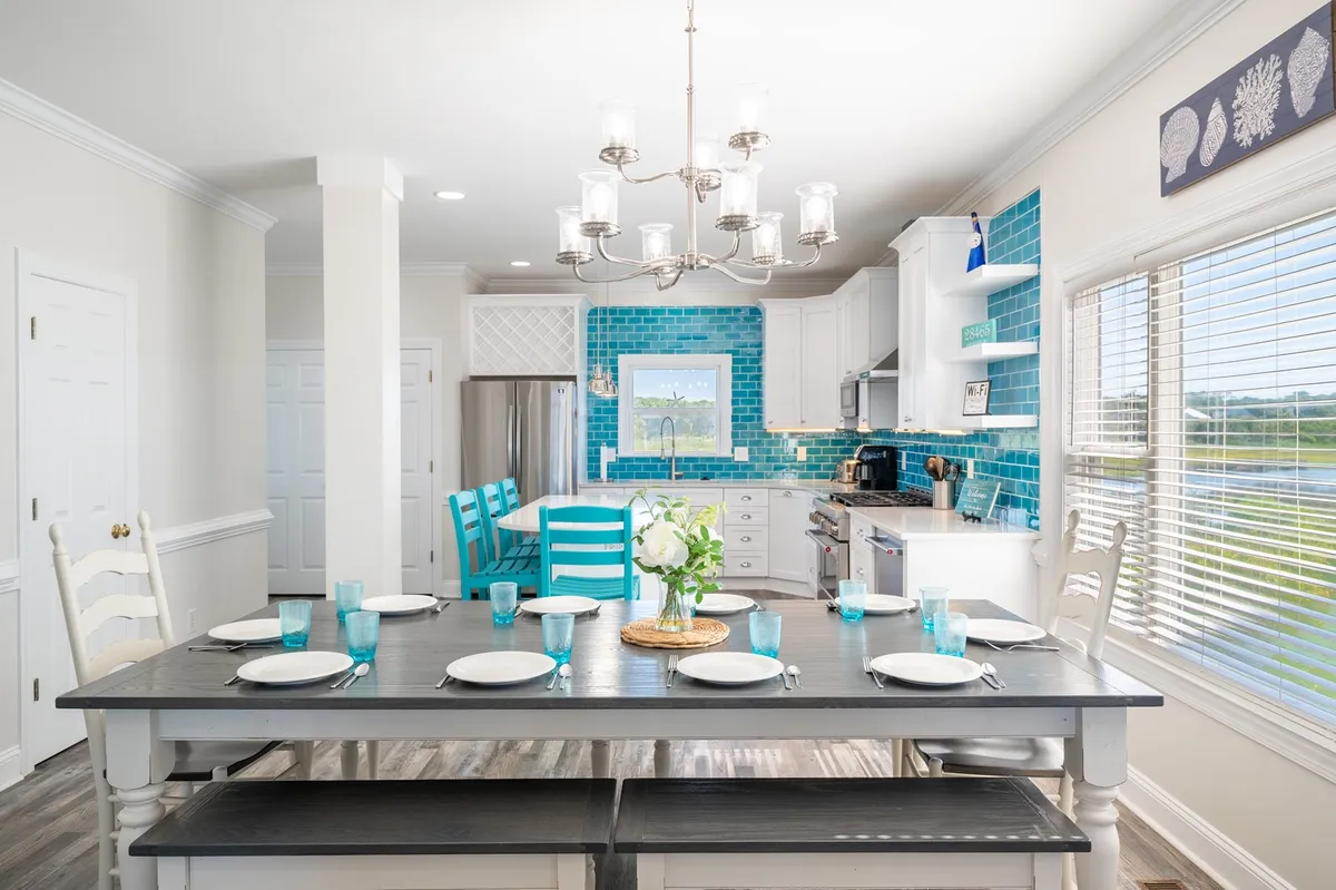 A spacious dining area with a large table under a striking chandelier, turquoise backsplash in the background, and a window view—offering a glimpse of sunny, relaxed gatherings.
