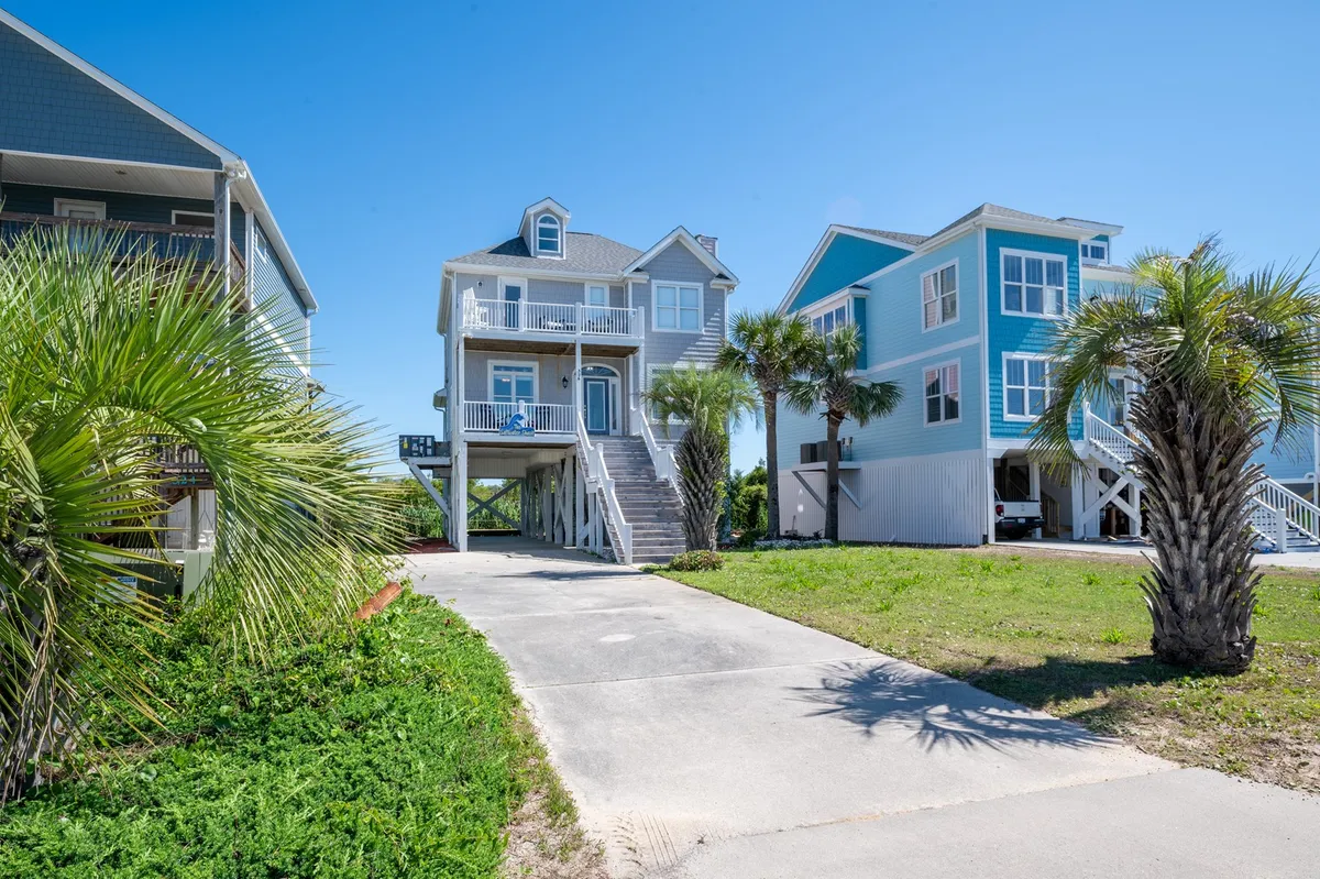 A cheerful view of the two-story beach house surrounded by palm trees and a welcoming driveway. The porch is visible, hinting at opportunities for relaxation or group activities in the fresh coastal air, and the overall neighborhood setting conveys the vibe of a retreat designed for friends and families.