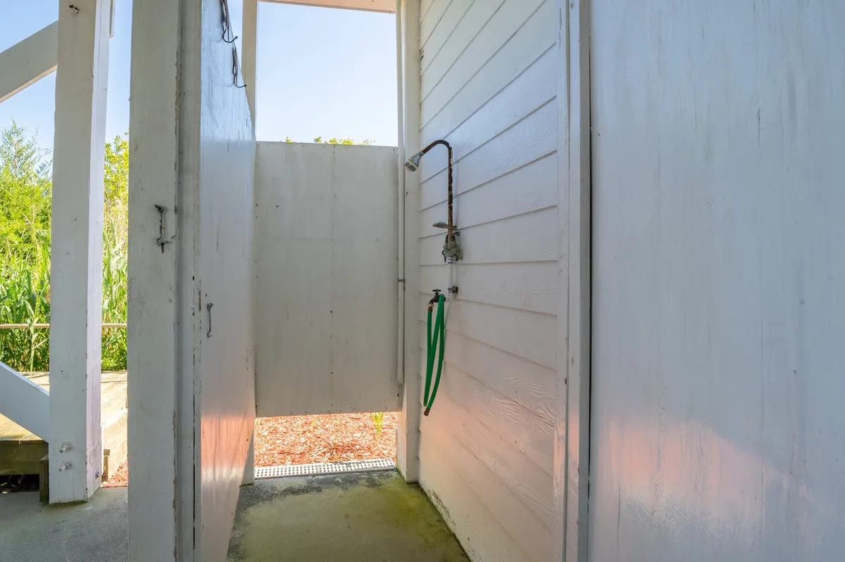 An inviting outdoor shower surrounded by greenery, with a garden hose and an open door leading to a natural area. The setting is private and refreshing, ideal for rinsing off sand after a day at the beach.