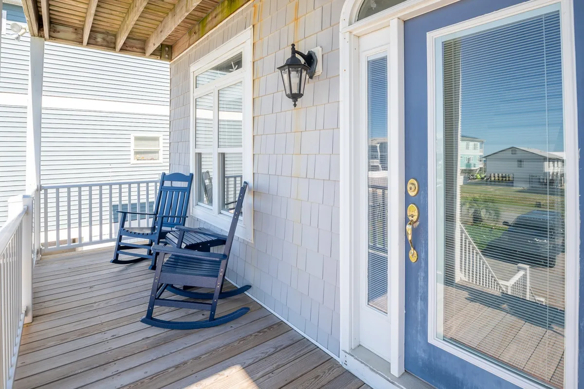 A charming front porch decorated with classic rocking chairs, a bright blue door, and windows that offer a glimpse of the interior. The scene is bathed in natural light, evoking a tranquil, welcoming atmosphere perfect for enjoying the outdoors.