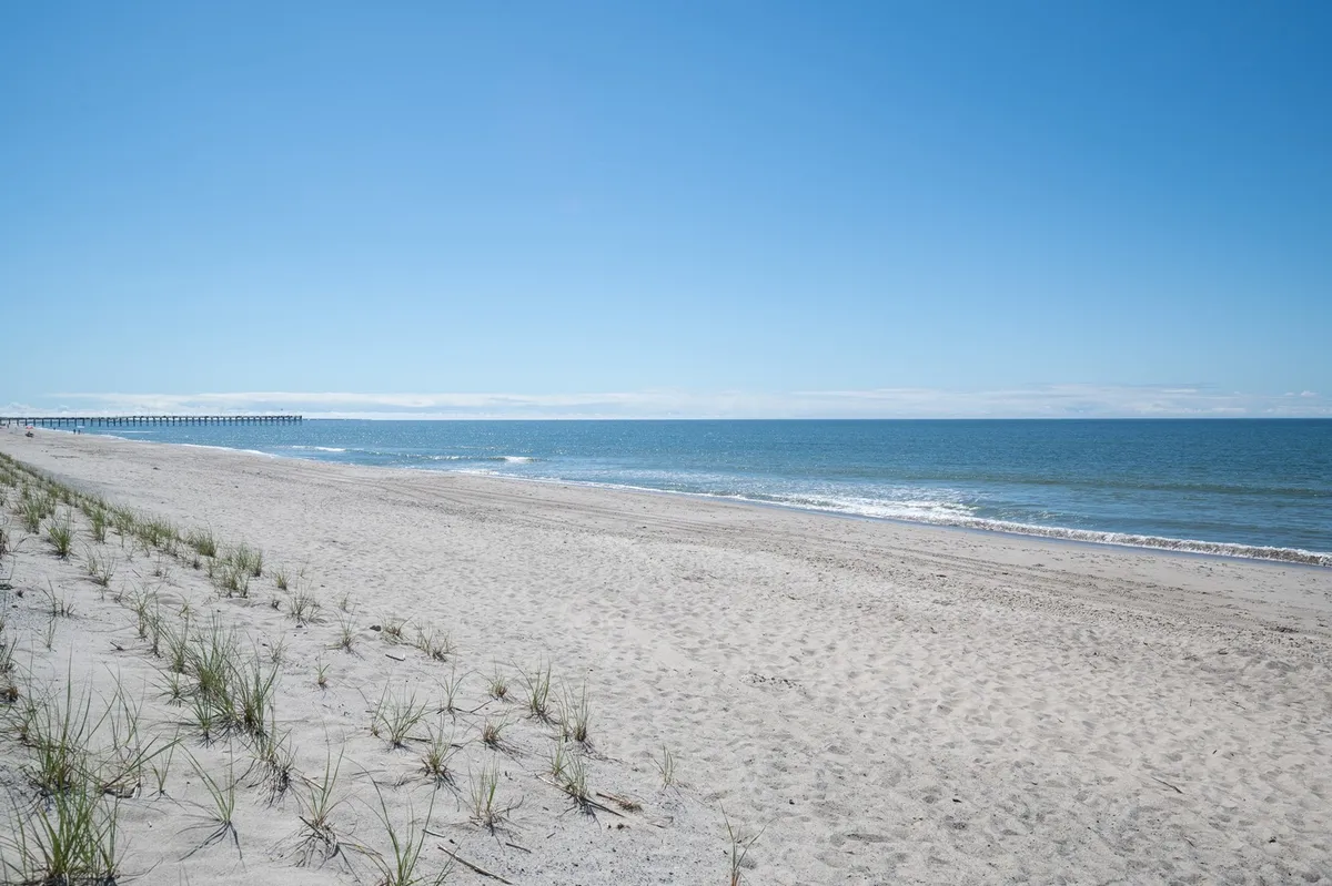 A sweeping view of Oak Island's sandy beach with rolling ocean waves, a wooden pier in the distance, and dune grass in the foreground. The scene is tranquil but lively, capturing both the pristine sand and the inviting water.