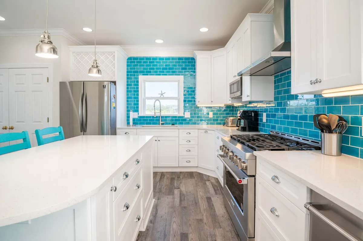 A modern, gourmet kitchen with a striking turquoise tile backsplash, substantial kitchen island, and high-end stainless steel appliances. Ample light fills the space, emphasizing the Italian tile and quartz countertops.