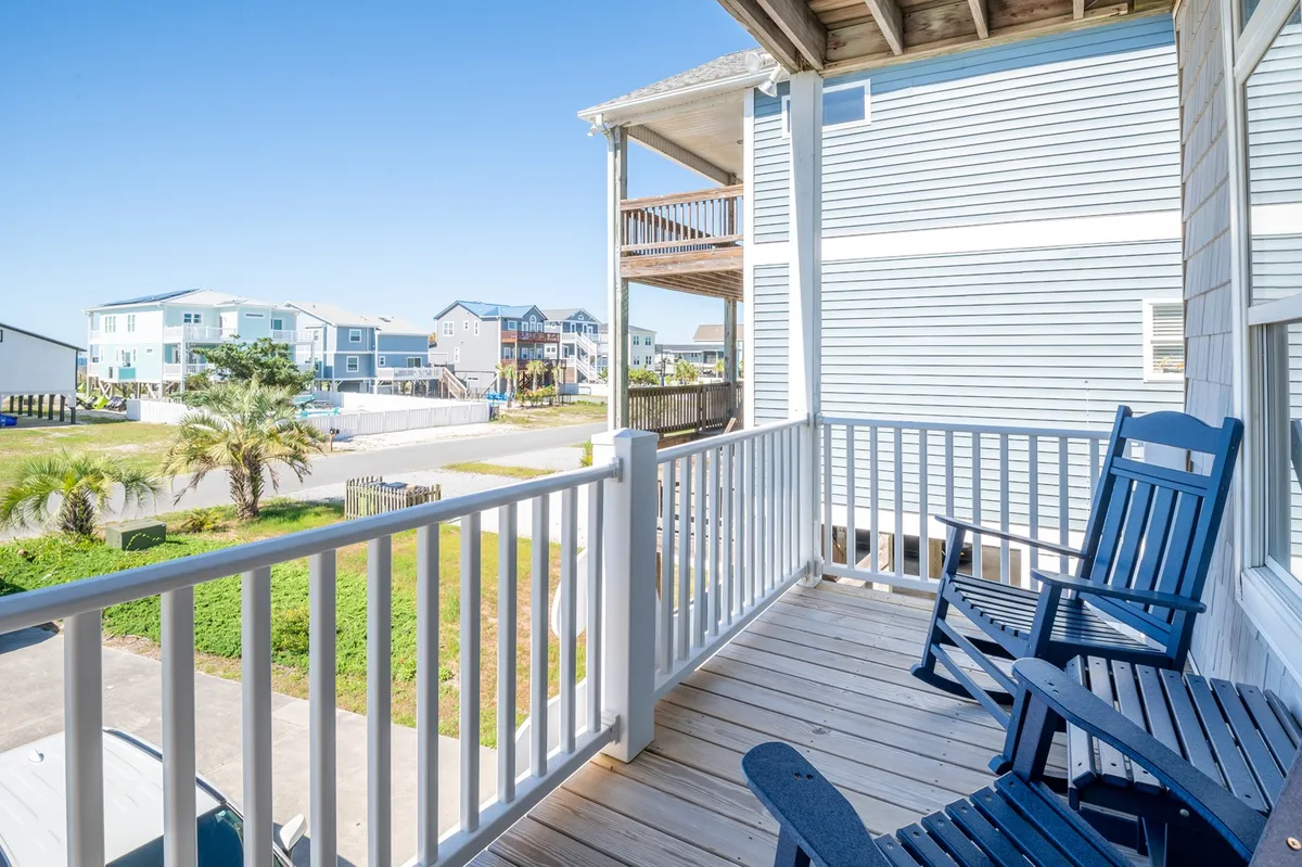 A spacious balcony featuring classic rocking chairs overlooks neighboring houses and well-maintained landscaping, inviting guests to linger and enjoy the open air. The arrangement of seating suggests a perfect spot for morning coffee or socializing with friends and family while watching the horizon.