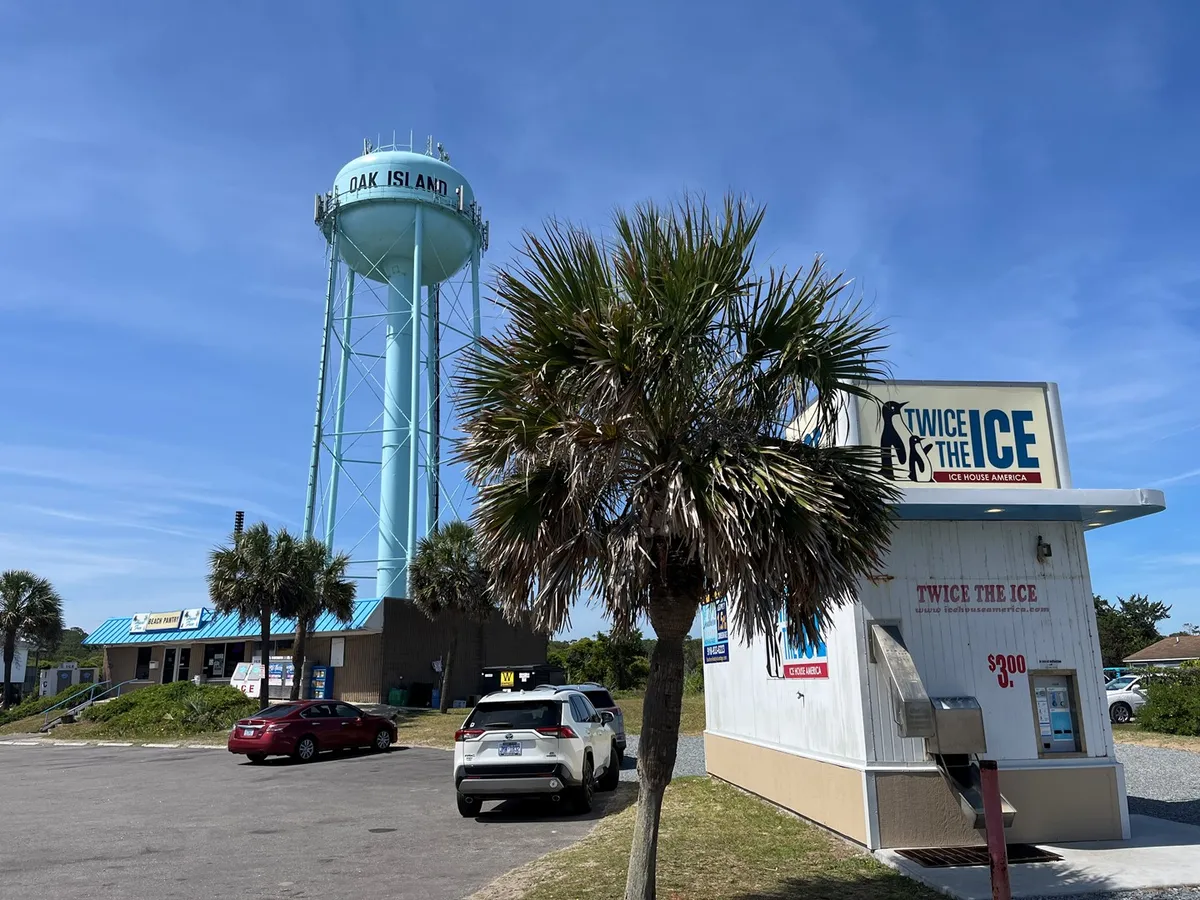 A relaxed community area featuring the iconic Oak Island water tower, palm trees, and a popular ice kiosk. The presence of cars and a casual gathering space evoke the fun, social energy of local events and community festivals.