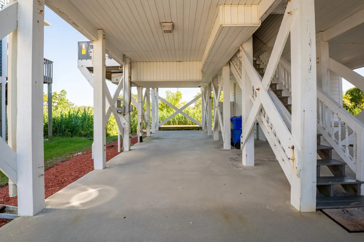 A spacious covered area beneath the house, supported by sturdy beams with a concrete floor and a blue recycling bin. The space provides ample room for parking cars or boat trailers, and offers quick access to outdoor adventures. The casual, functional vibe fits perfectly with the practical amenities described.