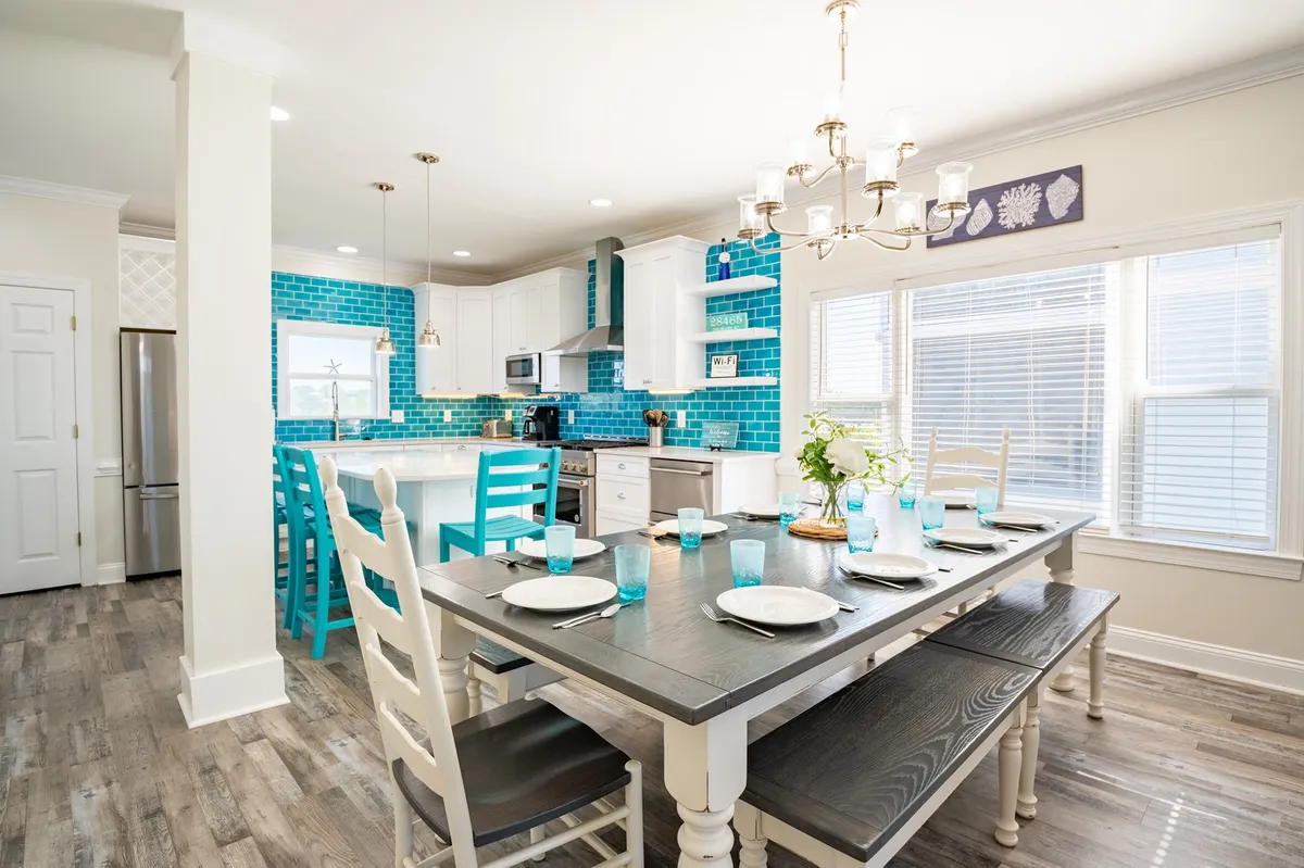 A cheerful, sunlit dining area featuring a large custom table set beneath a sparkling chandelier. The turquoise backsplash of the adjacent open kitchen and a spacious island are visible, hinting at gourmet amenities and inviting family gatherings.