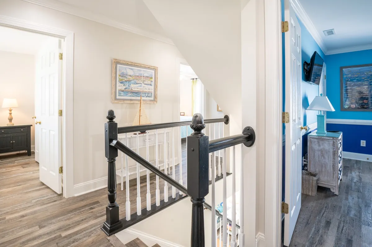 A bright hallway with a staircase railing, framed artwork, and a lamp on a blue wall, evoking the experience of entering or exiting the home—moments when guests are about to embark on (or return from) island excursions.