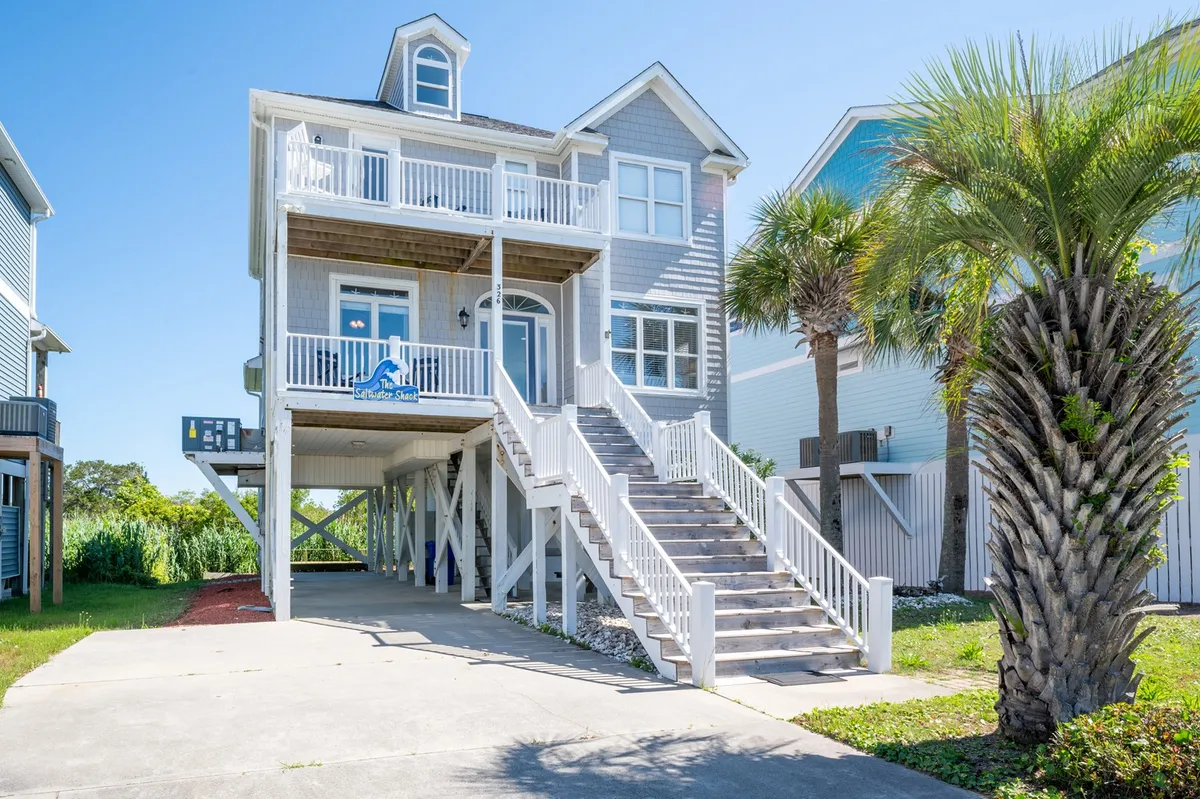 The image showcases the exterior of a beautifully remodeled beach house, with inviting balconies overlooking lush palm trees, a sunlit staircase leading up to the main entrance, and plenty of space for relaxing or gathering outdoors. The ocean view is visible just beyond the house, and the cheerful ambiance suggests a setting perfect for family vacations and sunset moments.