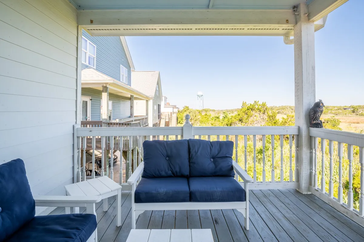 A cozy blue couch sits on an exterior porch with wooden railings, surrounded by lush greenery and offering distant views—including a water tower—across the neighborhood. The relaxed arrangement suggests a comfortable, private retreat for reading, chatting, or enjoying nature.